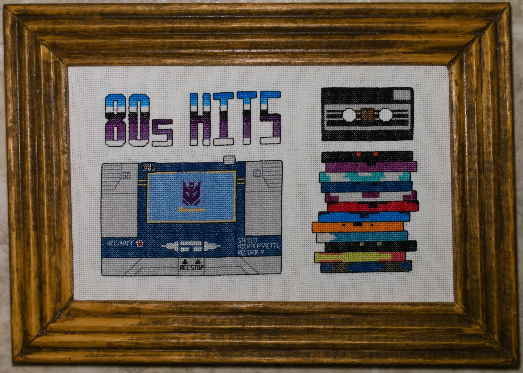 Framed Finished Cross Stitch Framed cross stitch. It contains a tape deck alt-mode Soundwave that sits under the words "80s HITS". Next to him sits a stack of cassttes, and above that stack is a cassette (Ravage) that is viewed from the front.
